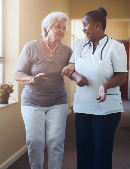 Portrait of happy healthcare worker walking and talking with senior woman. Elder woman gets help from nurse for a walk through nursing home.